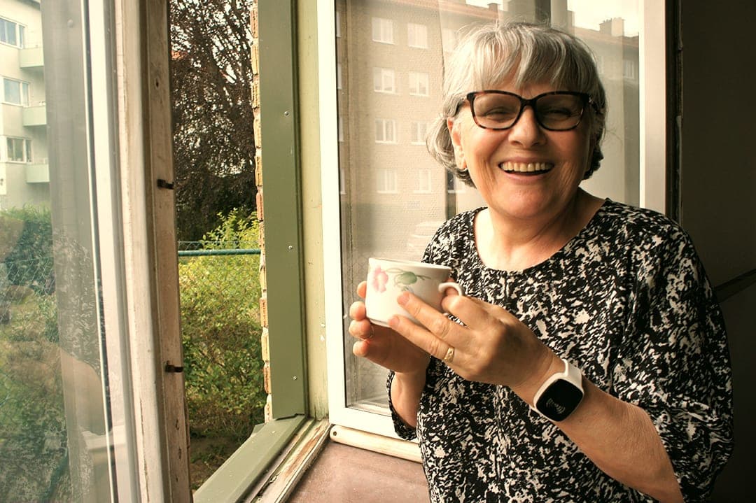 A smiling woman wearing TM-Alert on her wrist while holding a cup by an open window