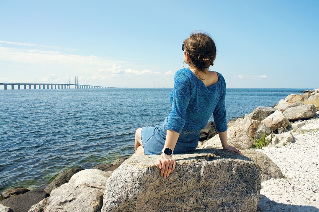 Young woman wearing a blue patterned top and denim skirt sitting on a large rock, gazing at the sea and a distant bridge on a bright sunny day, wearing a white TM-Alert wristband.