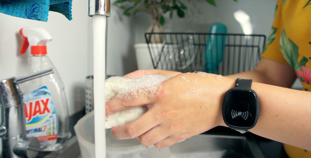 Close-up de uma pessoa lavando louça na pia da cozinha com água corrente e espuma de sabão nas mãos, enquanto usa uma pulseira inteligente TM-Alert preta.