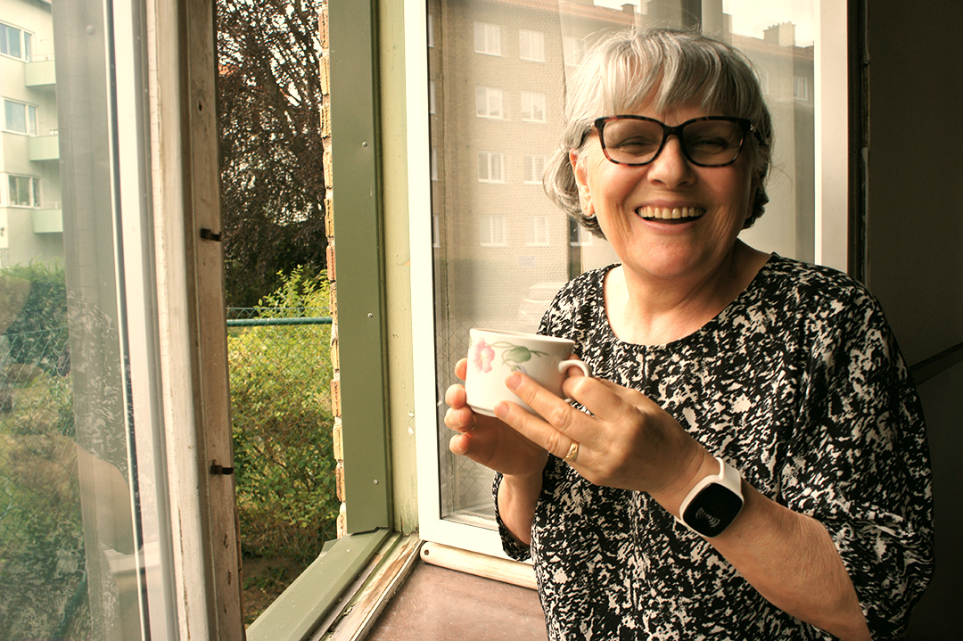 Elderly woman with gray hair and glasses smiling warmly while holding a floral mug by an open window. She wears a patterned black-and-white blouse and a white smart wristband with a black screen displaying a signal icon, suggesting an alert or assistive device. Residential buildings and greenery are visible outside.