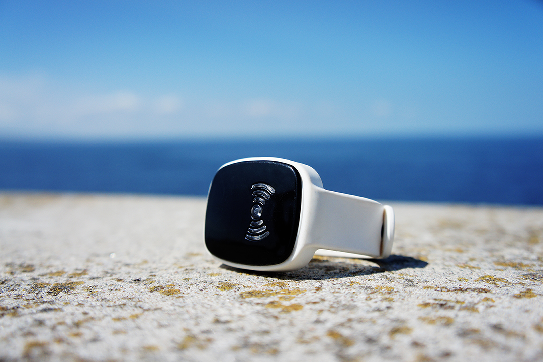 Close-up of a white smart wristband placed on a flat stone surface near the ocean, with the black screen facing forward and showing a subtle raised signal symbol. The blurred sea and sky in the background emphasize the product’s clean design and outdoor durability.
