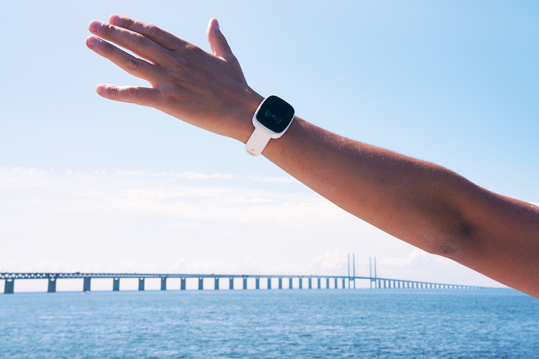 Outstretched arm of a person wearing a white wristband with a black digital screen displaying a green signal icon, raised against a clear blue sky and ocean with a long bridge in the background. Highlights visibility and connectivity of the wearable in open spaces.