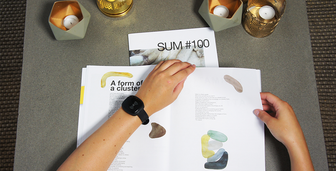 Overhead view of hands browsing an open design magazine with abstract art, wearing a black smart wristband. Decorative candles and the title “SUM #100” are on the table, showing the wearable’s presence in calm, indoor environments.