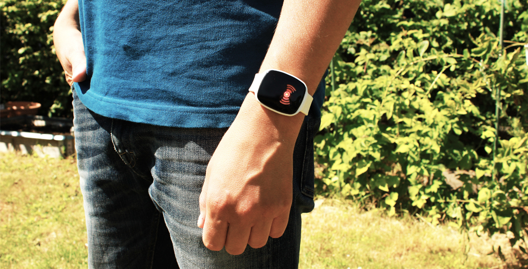 Person standing outdoors wearing a blue shirt and jeans, with a white smart wristband on their left wrist. The black screen of the device displays a glowing red alert icon, indicating an active warning or emergency feature.
